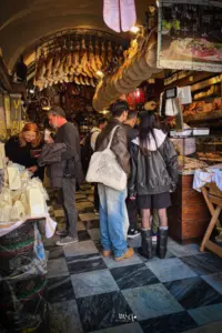 Einblick in eine typisch italienische Salumeria. Von der Decke hängen die Schinken und Salami Delikatessen und die Menschen stehne Schlange um etwas einzukaufen.
