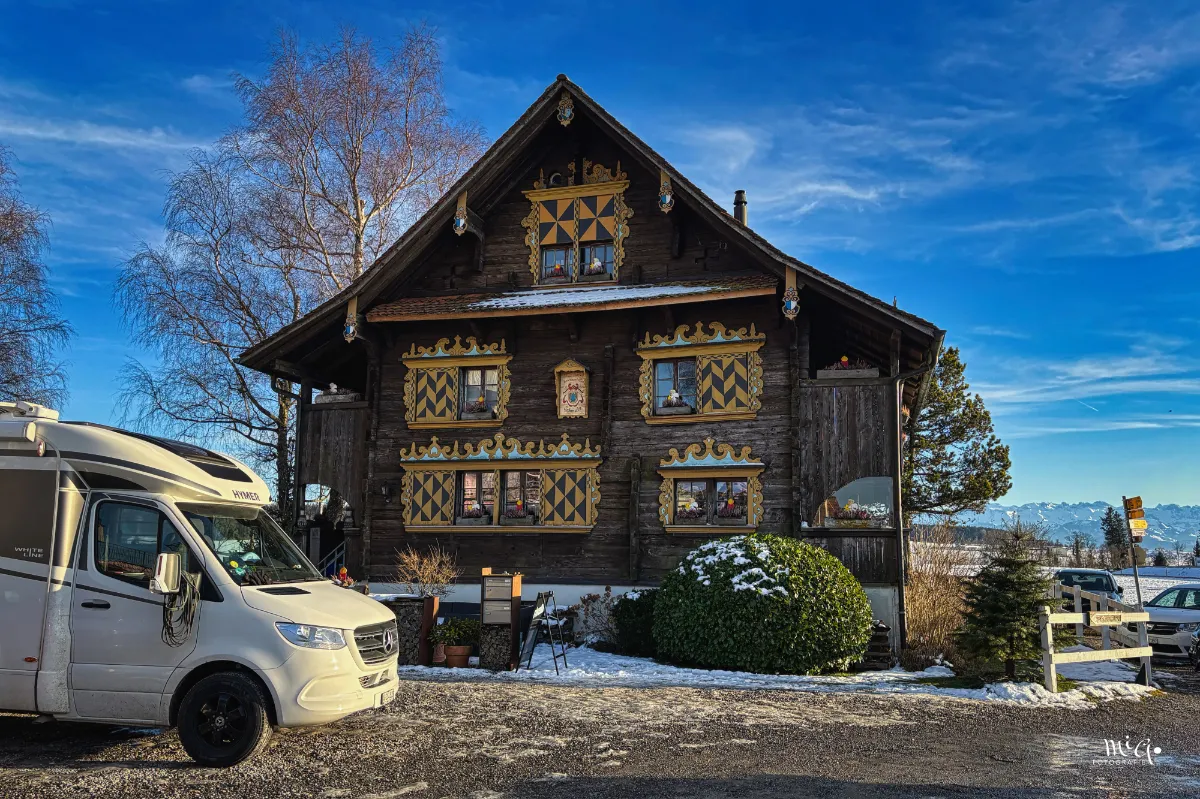Ein altes Holzhaus steht in der Landschaft mit einem davor parkierten Wohnmobil.