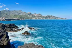 Blick über das tiefblaue Meer von Giardini Naxos nach Taormina das majestätisch auf den Hügeln über dem Meer thront.