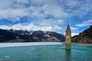 Das berühmte Bild, der aus dem Reschensee ragende Kirchturm. Der See ist zum Teil noch gefroren und es treiben Eisplatten im Wasser.