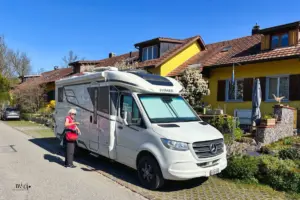 Eine Frau steht vor dem Wohnmobil welches vor einer Häuserreihe auf dem Parkplatz steht.